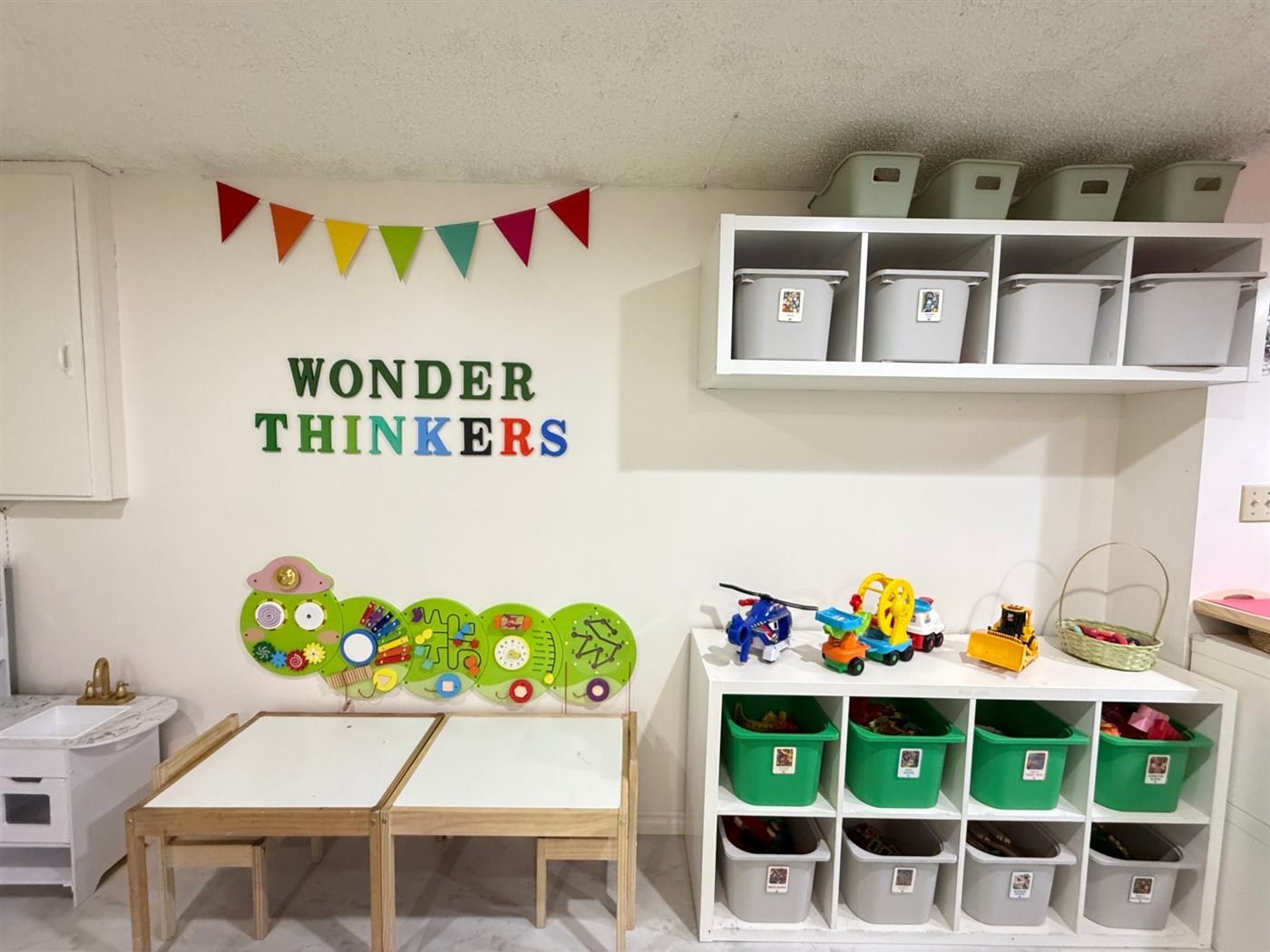 A green caterpillar busy-board on the wall above two low activity tables, with labelled toy bins and bunting beside it
