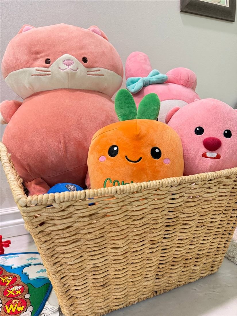 A woven basket filled with soft plush toys including a smiling carrot, a pink cat and a pink bunny